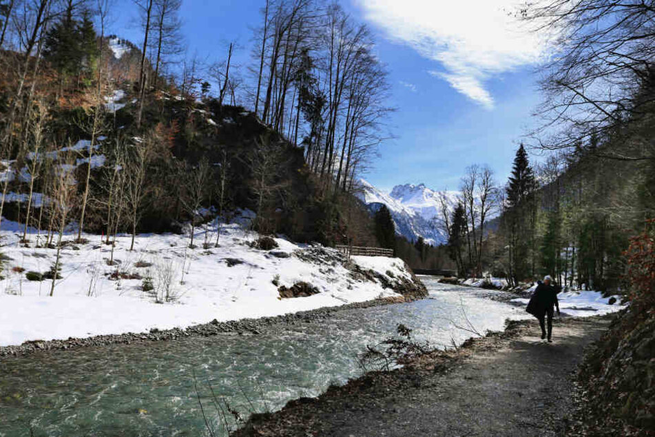 Eine Frau wandert entlang der Trettach bei Oberstdorf vor schneebedeckten Bergen im Sonnenschein.
