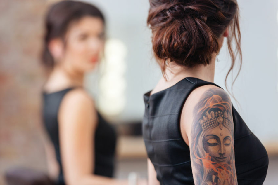 Eine Frau mit Tätowierungen sieht sich im Spiegel an. Claire Shepherd versteht nicht, warum Mitarbeiter nicht auch Tattoos tragen dürfen. (Symbolbild)