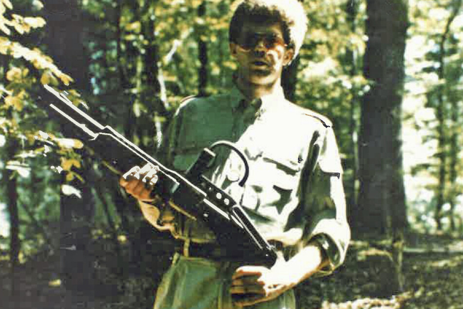 Das von der belgischen Polizei veröffentlichte Foto zeigt einen Mann mit Sonnenbrille, der im Wald steht, militärähnliche Kleidung trägt und eine Waffe hält.