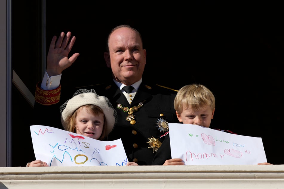 Fürst Albert II. von Monaco (63) winkt während der Feierlichkeiten zum Nationalfeiertag vom Balkon des Palastes von Monaco, während seine Kinder Fürst Jacques und Prinzessin Gabriella von Monaco (beide 7) Bilder mit Botschaften für ihre Mutter in der Hand halten.