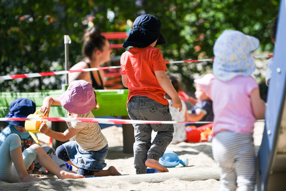Kinder einer Krippen-Gruppe spielen auf dem Spielplatz der Kita. Absperrbänder regeln den Abstand.