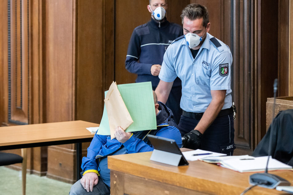 Einer der beiden Angeklagten wird im Rollstuhl und mit einer Mappe vor dem Gesicht vor Beginn der Verhandlung in den Gerichtssaal des Landgerichts geschoben.