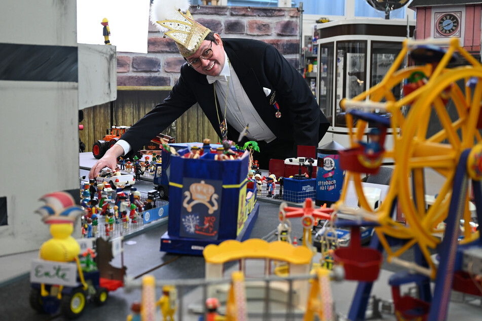 Simon Isser, Sitzungspräsident des Offenbacher Karnevalvereins (OKV), steht in einer Halle in Mühlheim hinter einen nachgebauten Kulisse mit bunten Miniatur-Motivwagen, in denen Playmobilfiguren platziert sind.