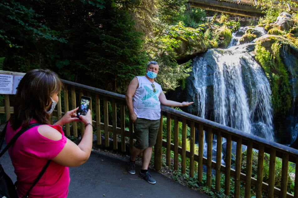 Ein Tourist steht an einem Geländer und posiert für ein Foto, während seine Begleiterin mit ihrem Smartphone ein Bild von ihm vor einer Stufe der Triberger Wasserfälle macht.