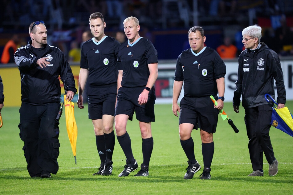 Schwerstarbeit für das Schiedsrichter-Gespann bei einem Spiel von Lok Leipzig. Zwei Vereinsmitarbeiter haben die obligatorischen Regenschirme zur Abwehr von Wurfgeschossen dabei.