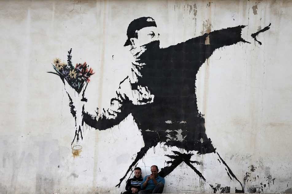 Two men sitting in front of famous graffiti by British street artist Banksy, painted on a wall of a gas station in the West Bank city of Bethlehem in December 2015.