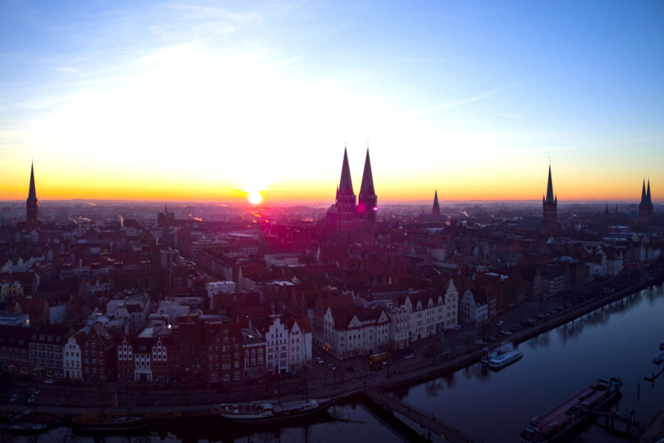 Die Sonne geht über die Hansestadt, der Stadt der sieben Türme, auf: Kirche St. Jakobi (v.l), Marienkirche, die drei Spitzen des Rathauses, St.-Aegidien-Kirche, Petrikirche und der Dom zu Lübeck. Im Vordergrund fließt die Trave.