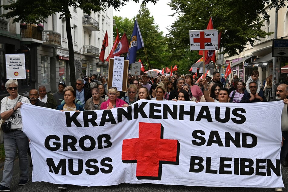 Demonstranten halten ein Banner mit der Aufschrift "Krankenhaus Groß-Sand muss bleiben" in die Luft.