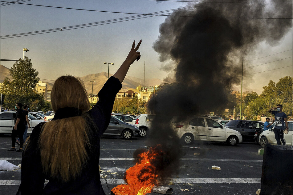 Eine Frau steht während einer Demonstration nach dem Tod von Mahsa Amini (†22) vor einem brennenden Autoreifen und zeigt das Friedenszeichen.