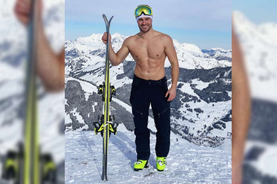 Alexander Hindersmann posiert mit freiem Oberkörper und Skiern in der Hand vor einer beachtlichen Bergkulisse.