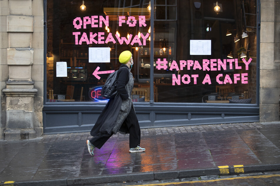 Eine Frau geht auf der Cockburn Street am Bistro "Scran" vorbei, auf dessen Fensterscheiben "Open For Takeaway" (Für Essen zum Mitnehmen geöffnet) und der Hashtag "Apparently Not a Cafe" (Anscheinend kein Café) steht.