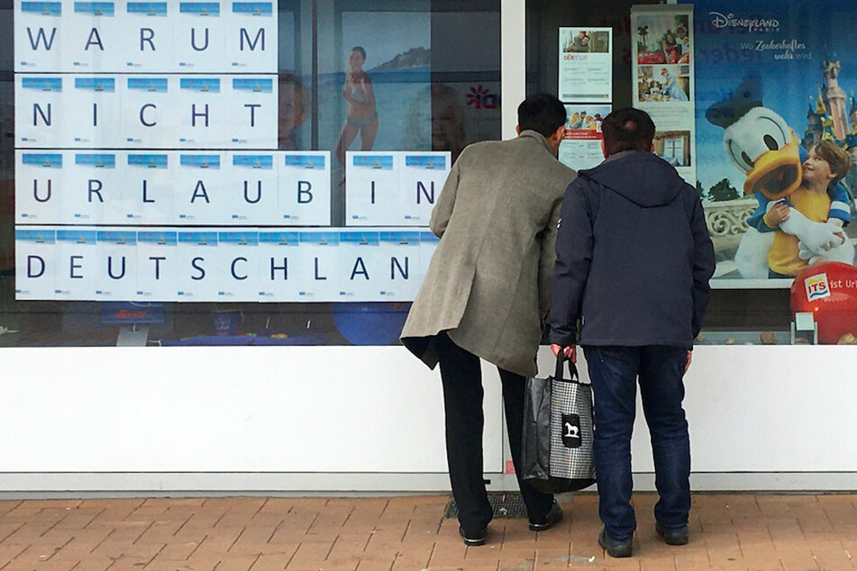Zwei Männer schauen in das Schaufenster eines Reisebüros, an dem mit großen Buchstaben "Warum nicht Urlaub in Deutschland" zu lesen ist.
