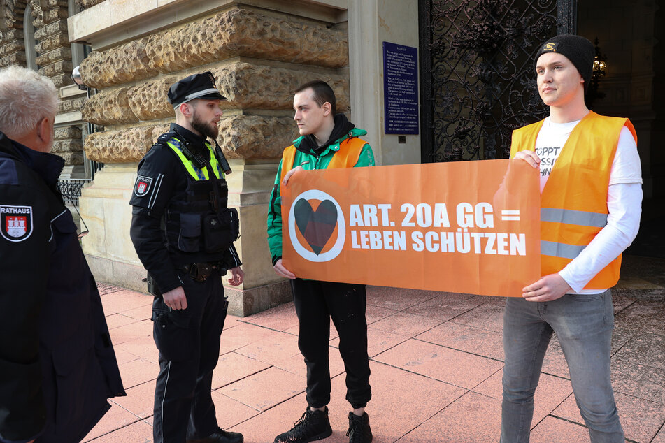 Klimaaktivisten der Bewegung Letzte Generation, die zuvor das Hamburger Rathaus besprüht haben, stehen mit einem Transparent mit der Aufschrift "Art. 20A GG = Leben Schützen" vor dem Rathauseingang.
