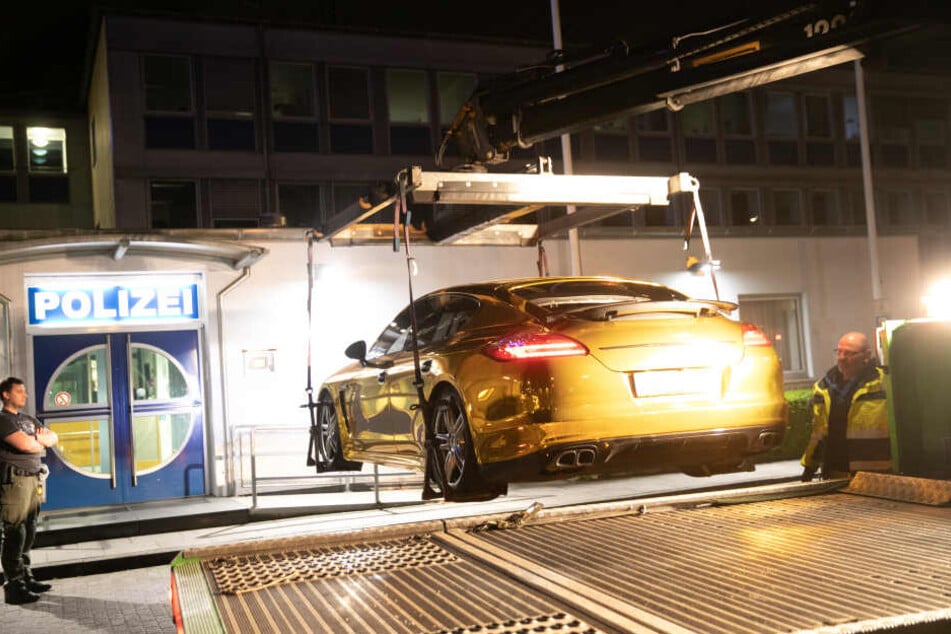 Ein Polizist steht vor einer Polizeiwache an einem mit Goldfolie beklebten Porsche Panamera, der auf einen Fahrzeugtransporter verladen wird.