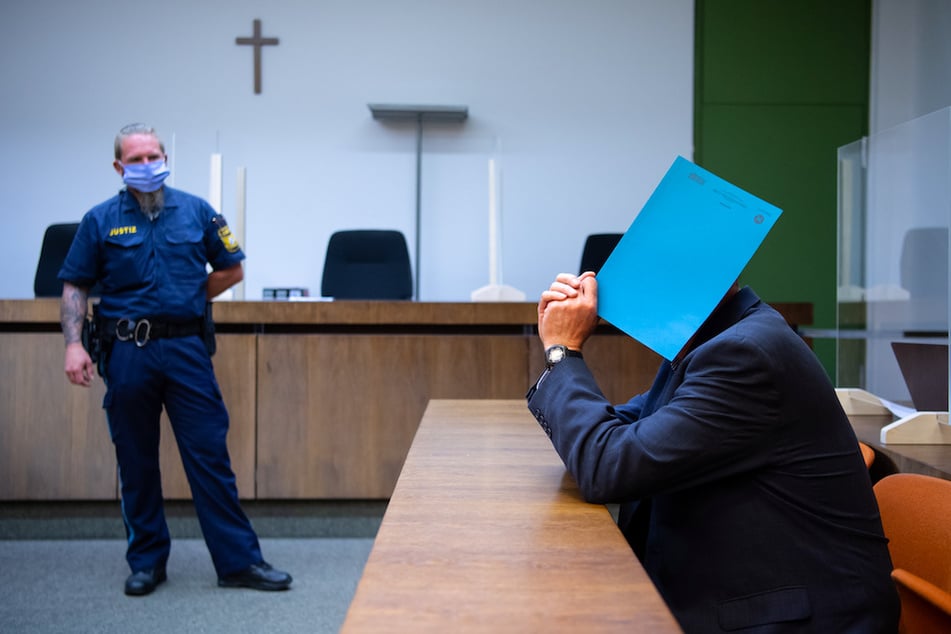 Der wegen sexuellen Missbrauchs von Kindern und Schutzbefohlenen sowie sexueller Nötigung angeklagte Mann (r) sitzt vor Prozessbeginn im Landgericht auf seinem Platz und hält sich einen Ordner vor das Gesicht.