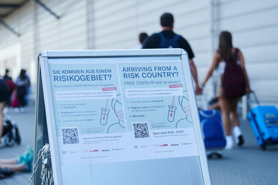 "Sie kommen aus einem Risikogebiet?" und weitere Informationen stehen auf einem Schild am Flughafen Schönefeld, während Passagiere mit Rollkoffern vorbeilaufen.