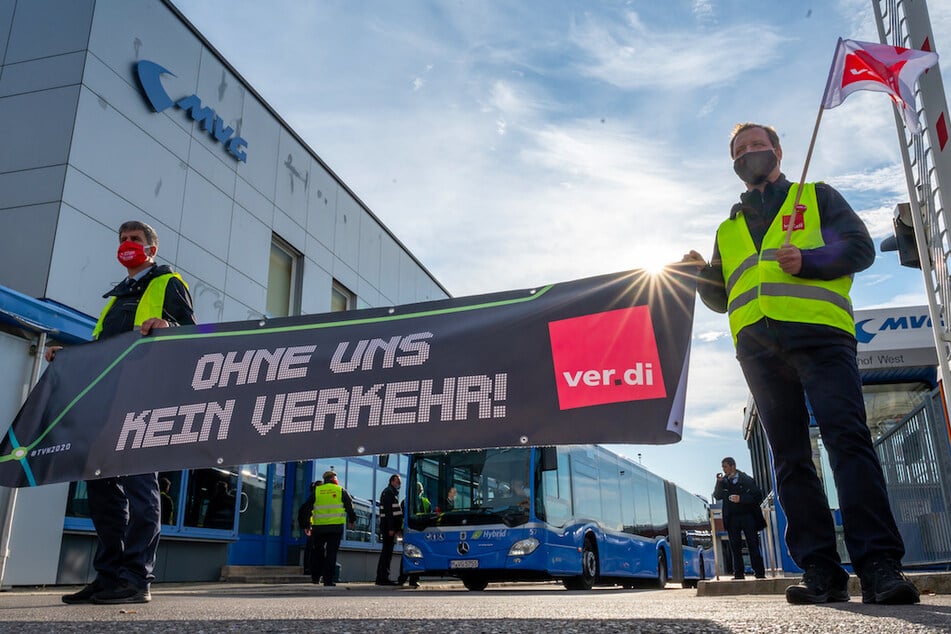 Mitarbeiter der Münchner Verkehrsgesellschaft (MVG) stehen mit Verdi-Fahnen und einem Banner mit der Aufschrift "Ohne uns kein Verkehr" vor einem Omnibusbahnhof. (Archivbild)