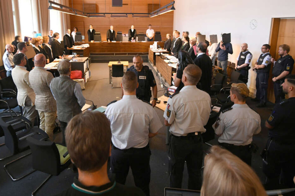 Zu Beginn des Prozesses stehen elf Angeklagte und Justizbeamte im Landgericht seitlich hinter ihren Anwälten. In der Mitte stehen die Richter und Schöffen. (Archivbild)