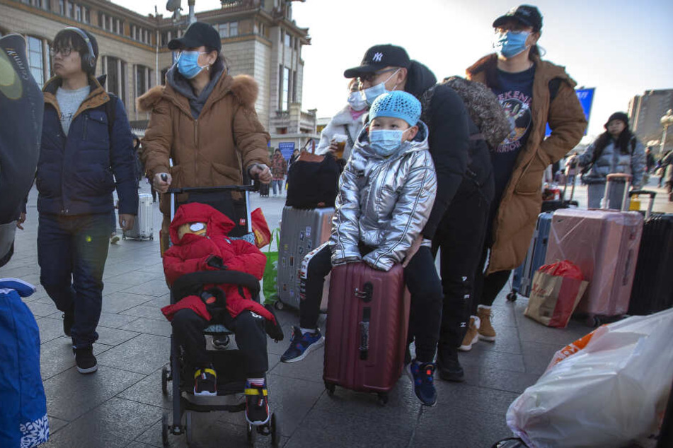 Reisende stehen mit Rollkoffern in der Nähe des Pekinger Bahnhofs und tragen Mundschutz.