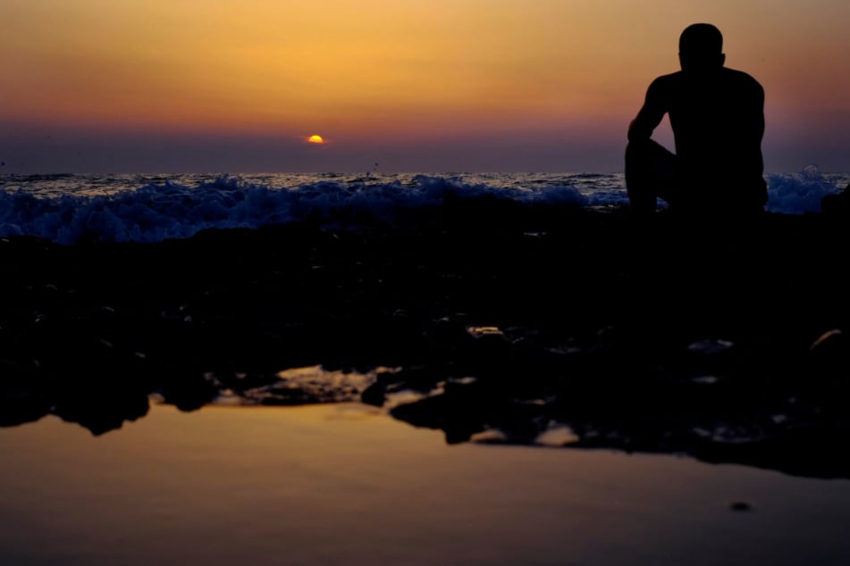 Zypern, Ayia Eirini: Ein Mann sitzt bei Sonnenuntergang auf einem Felsen am Meer.
