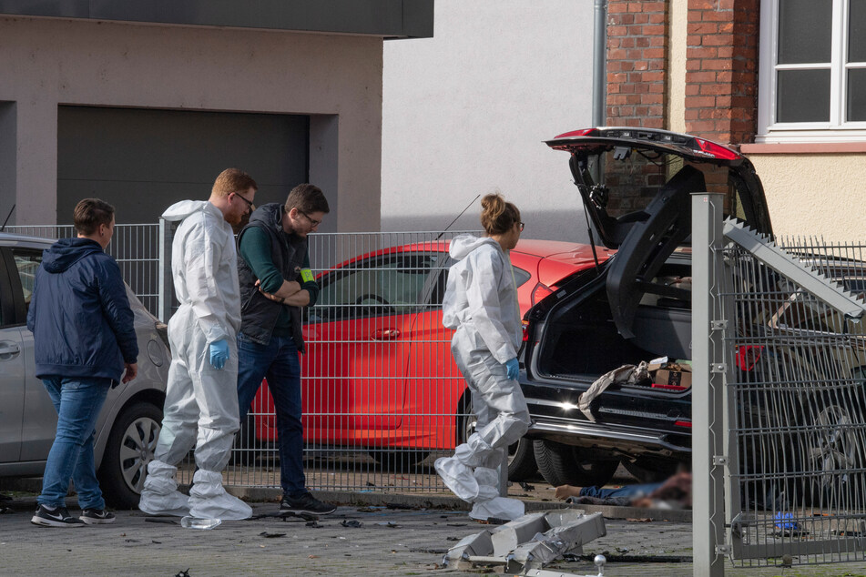 Mitarbeiter der Spurensicherung stehen in der Innenstadt an dem Wrack eines dunklen Pkw, unter dem die Leiche einer Frau liegt.