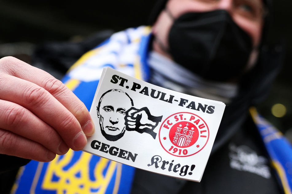 Ein Fan des FC St. Pauli trägt einen Schal in den Farben der Ukraine und einen Aufkleber mit dem Text "St. Pauli-Fans gegen Krieg".