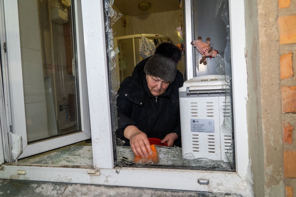 Eine Bewohnerin des Nachbarhauses kehrt nach einem russischen Luftangriff zerbrochenes Fensterglas zusammen.