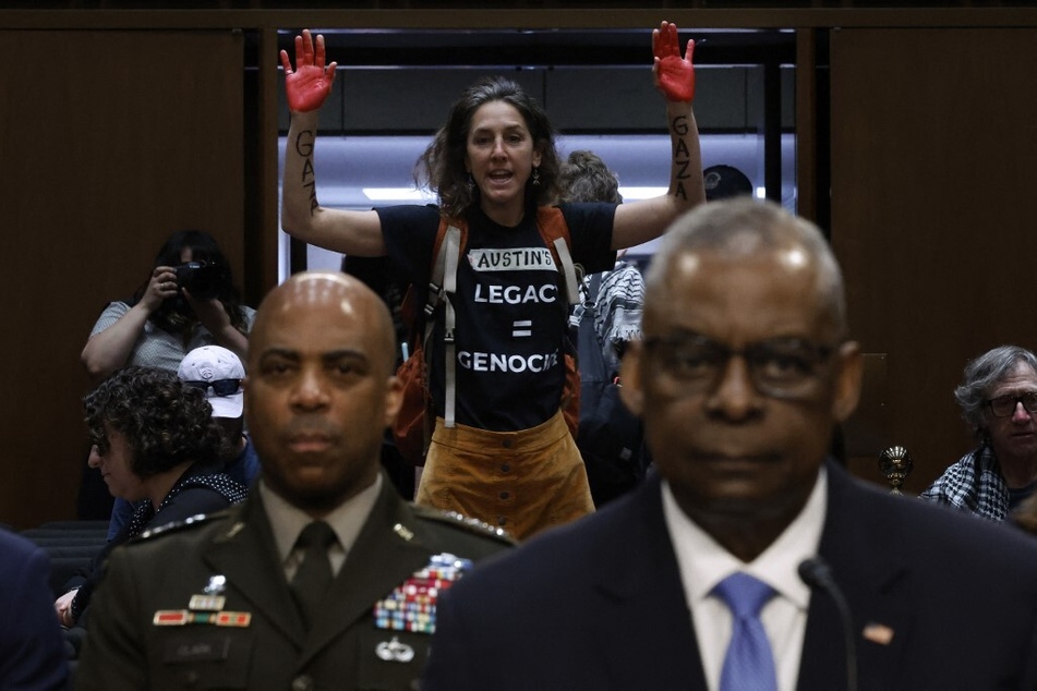 A protester with red-painted hands and a shirt reading "Austin's Legacy = Genocide" disrupts Secretary of Defense Lloyd Austin's testimony before the Senate Armed Services Committee.