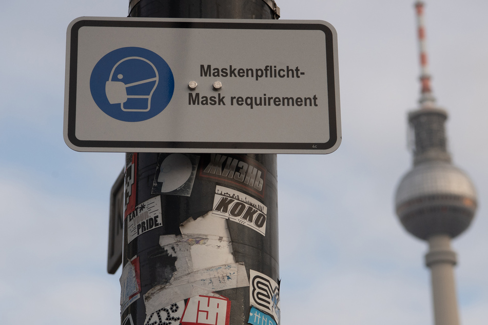 Berlin: Ein Hinweisschild auf die Maskenpflicht hängt an einem Mast, im Hintergrund der Fernsehturm.