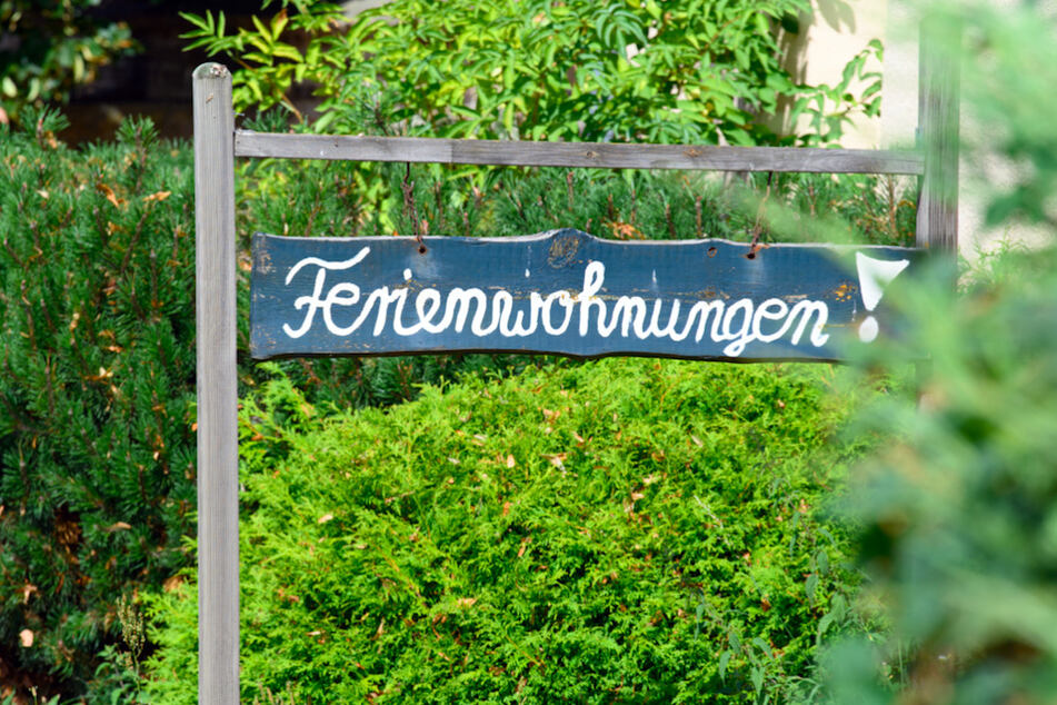 Ein Schild „Ferienwohnungen!“ steht vor einem Haus zwischen Bäumen und Sträuchern.