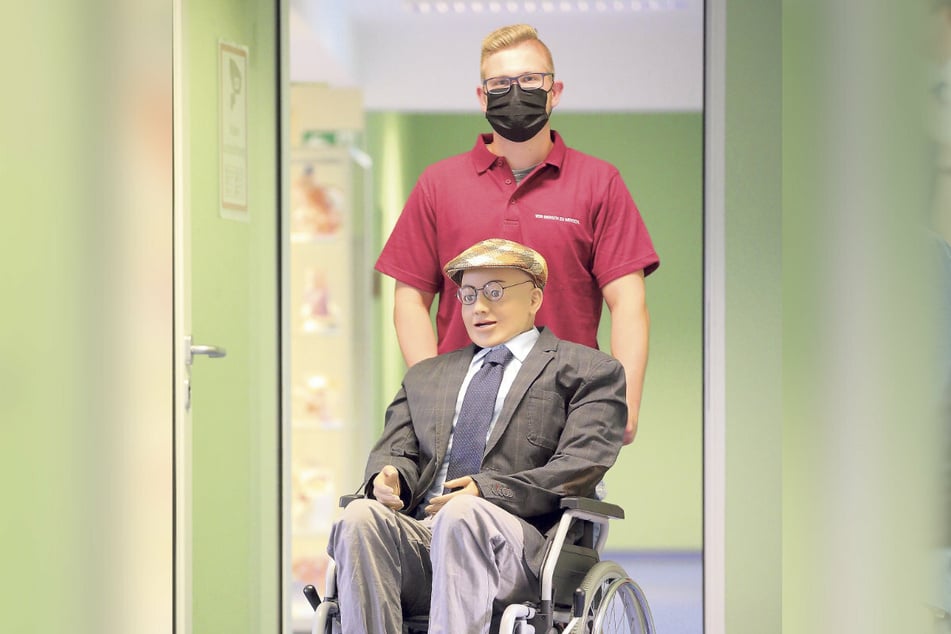 Pflegestudent Yannick Klaus (21) schiebt "Herrn Schuster" - der eigentlich eine technisch ausgefeilte Puppe ist - im Rollstuhl über den Gang des Simulationslabors.