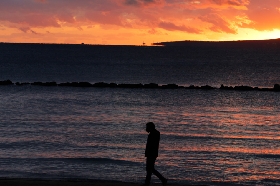 Ein Mann mit Gesichtsmaske spaziert während des Sonnenuntergangs am Strand von Palma entlang.