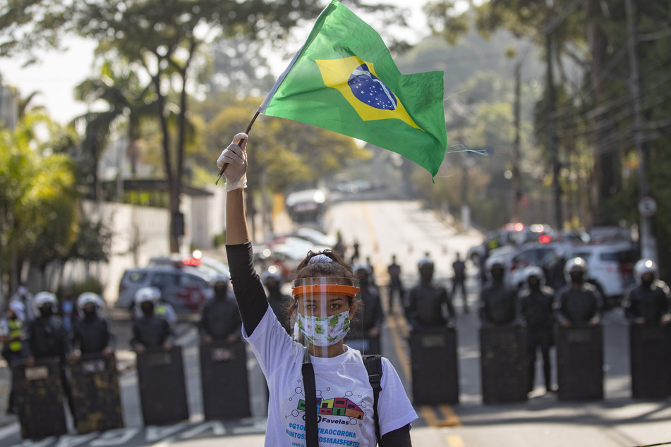Eine Bewohnerin der Favela Paraisopolis mit Mundschutz und Gesichtsmaske schwenkt eine brasilianische Fahne vor einer Reihe Polizisten während eines Protestes. Bewohner der Favela forderten stärkere Maßnahmen seitens des Staates gegen die Ausbreitung des Coronavirus.