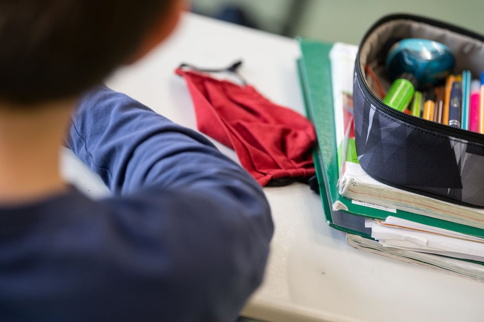 Eine Mund-Nasen-Bedeckung liegt während einer Unterrichtsstunde neben einem Mäppchen und Schulbüchern auf einem Tisch.