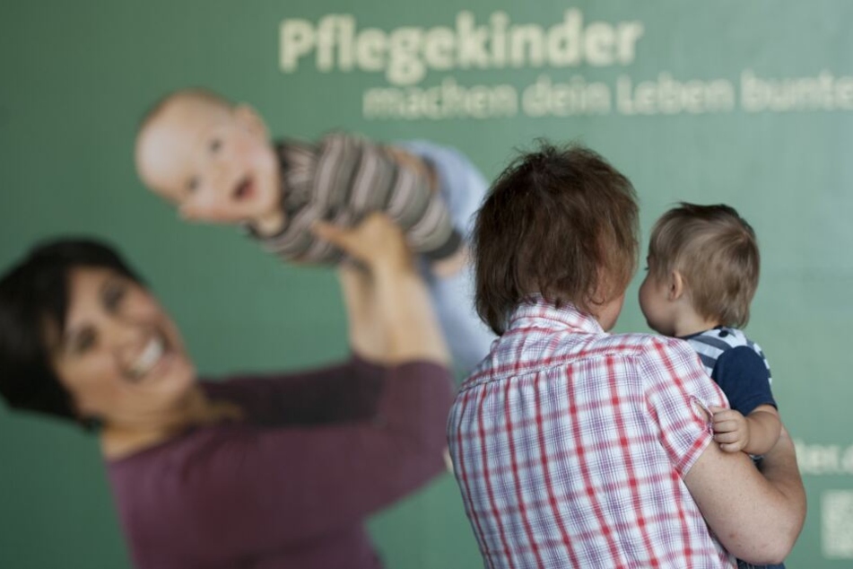 Eine Frau steht mit einem Kind im Arm vor einem Plakat mit der Aufschrift "Pflegekinder machen dein Leben bunter".