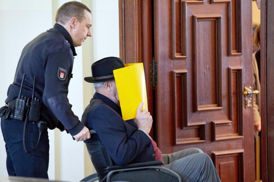 Der Angeklagte wird in einem Rollstuhl während einer Prozess-Pause aus den Gerichtssaal gebracht.