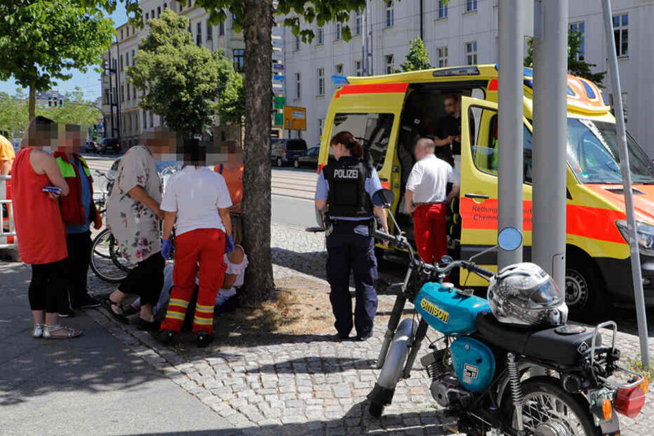 Vermutlich die Simson-Fahrerin lehnt hier sitzend an einem Baum. Passanten und Sanitäter kümmern sich um sie.