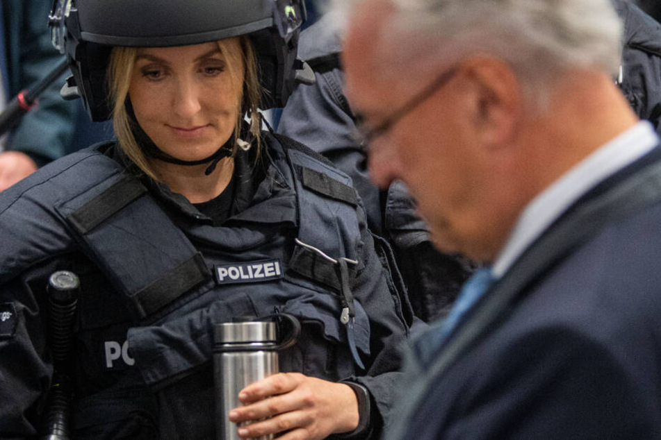 Eine Einsatzkraft der Bereitschaftspolizei bekommt eine Thermoskanne mit dem Shake ausgehändigt. Rechts steht Bayerns Innenminister Herrmann (CSU).