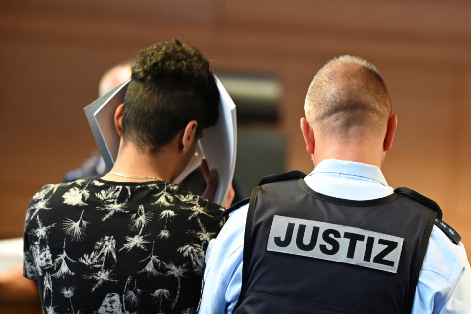 Einer von elf Angeklagten wird vor dem Prozessbeginn von einem Justizbeamten in einen Gerichtssaal im Landgericht gebracht. (Archivbild)