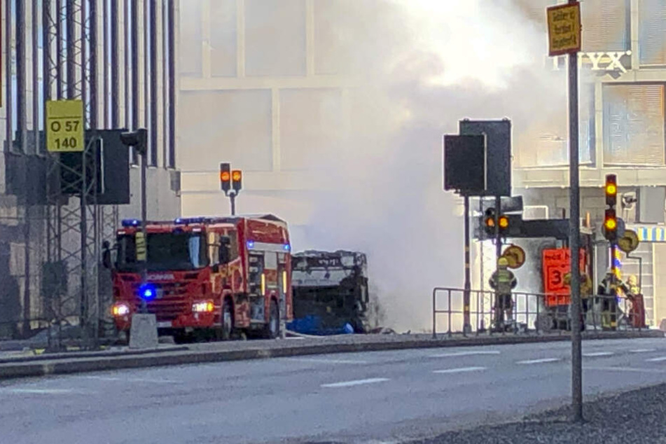 Einsatzkräfte der Feuerwehr stehen nach der Explosion eines Busses im Stadtzentrum neben dem ausgebrannten Bus.