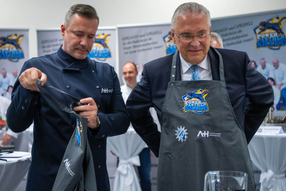 Joachim Herrmann (r, CSU), Innenminister von Bayern, zieht bei der Vorstellung der Shakes neben Alexander Herrmann, TV-Koch, eine Schürze an.
