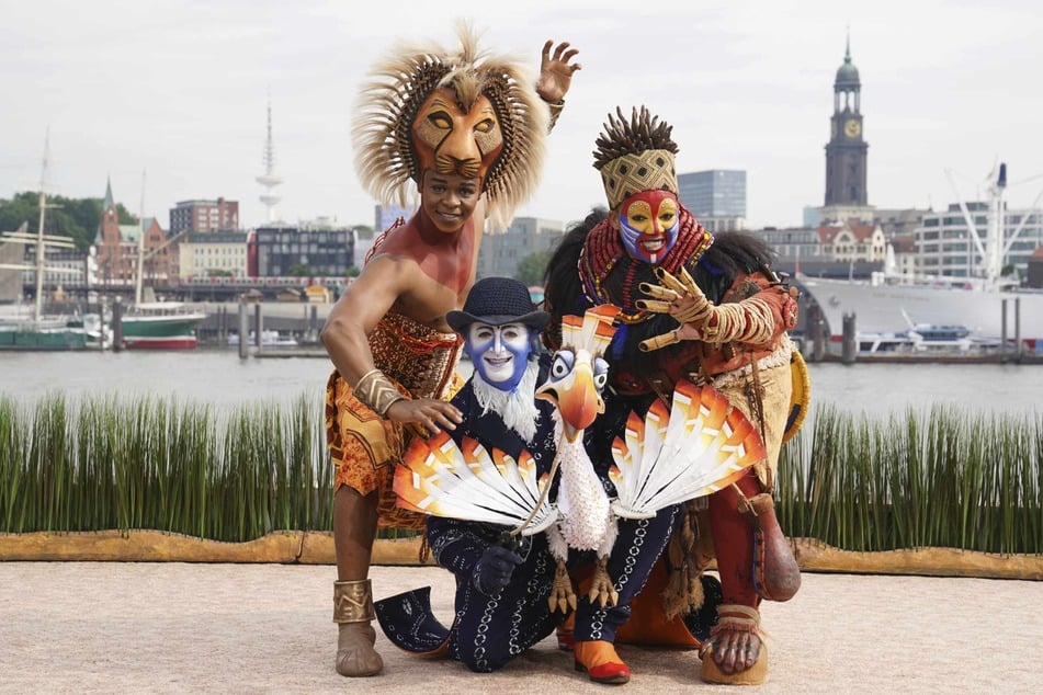 Die Musical-Darsteller aus "Der König der Löwen", Hope Maine als Simba (v.l.n.r.), Joachim Benoit als Zazu und Thenjiwe Nofemele als Rafiki, stehen auf einer kleinen Bühne vor dem Musicaltheater im Hafen und vor der Skyline der Hansestadt.