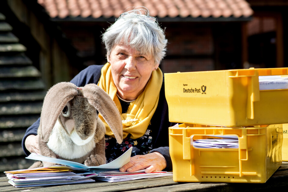 Doris Kröger, Mitarbeiterin des Osterpostamts, sitzt mit einem Osterhasen aus Stoff und vielen Briefen vor dem Gemeindehaus im Ortskern von Ostereistedt.