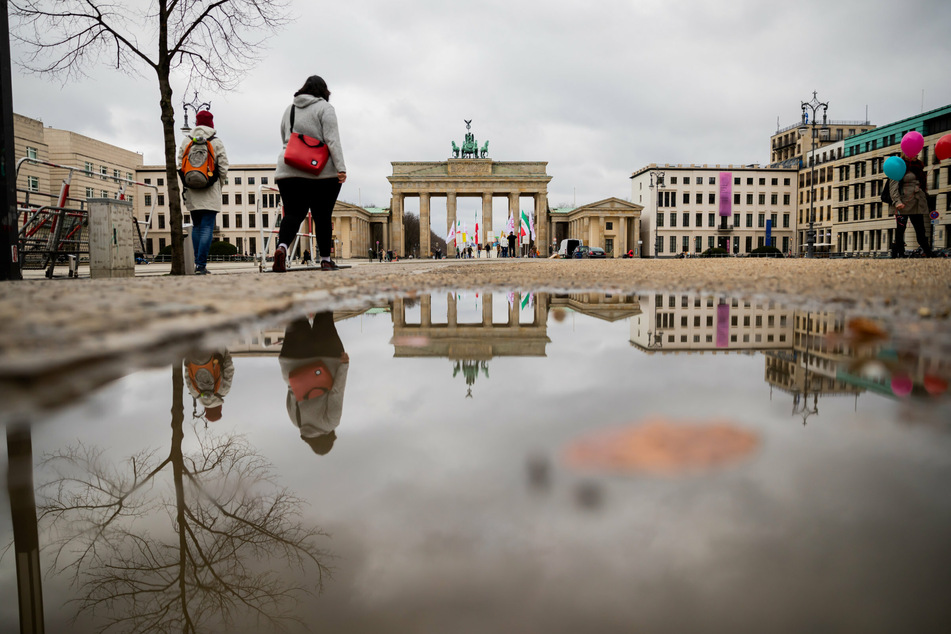 Passanten gehen vor dem Brandenburger Tor, welches sich bei bedecktem Himmel in einer Pfütze spiegelt.