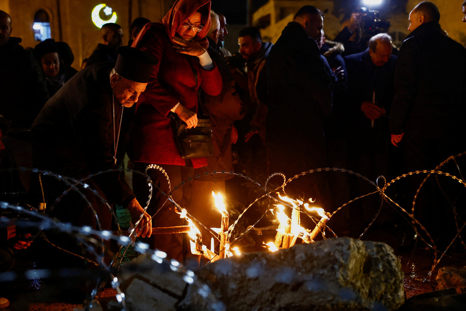 A priest lights a candle at a Christmas installation of a grotto with figures standing amid rubble surrounded by a razor wire outside the Church of the Nativity in Bethlehem, in the Israeli-occupied West Bank.