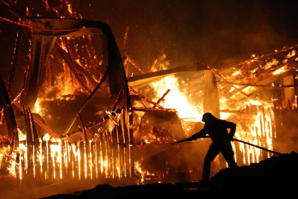 Eine Einsatzkraft der Feuerwehr steht vor dem lichterloh brennenden Stall in Rettenberg und kämpft gegen die Flammen.
