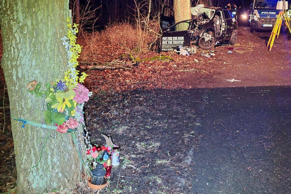 Im Hintergrund ist das Wrack des VW Polo zu sehen, im Vordergrund sieht man am nächsten Baum, an dem sich vor fünf Jahren augenscheinlich ein weiterer tödlicher Verkehrsunfall ereignet hat, Blumen und Kerzen stehen.