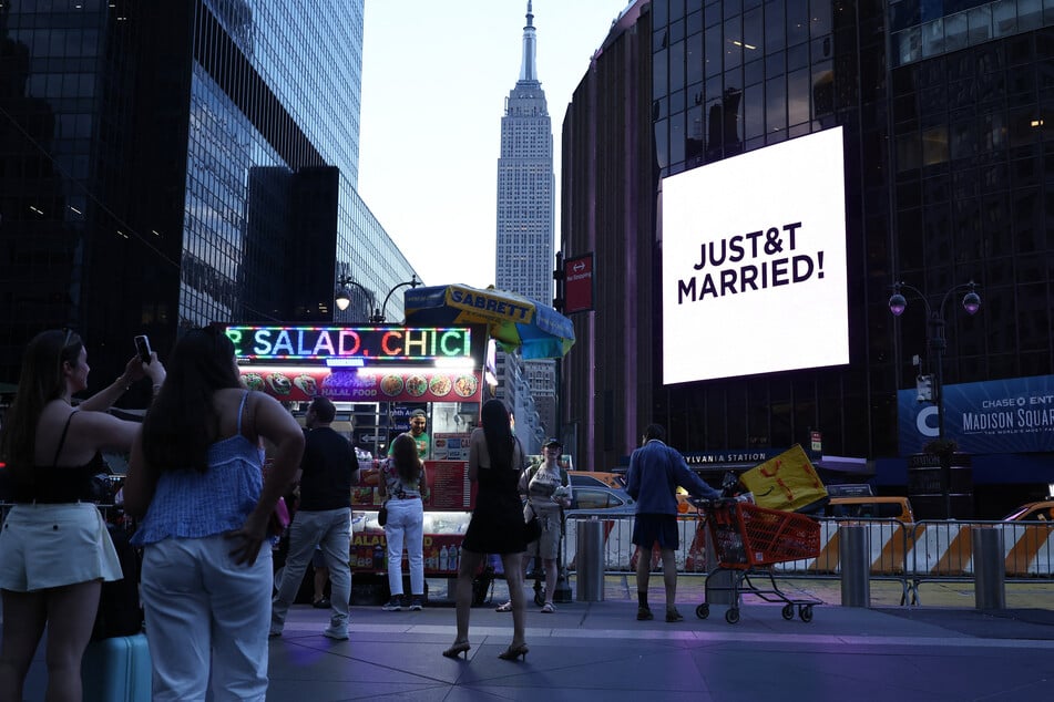 The Empire State Building is seen in the distance as people take photos of a giant screen showing the message "JUST&T MARRIED!"