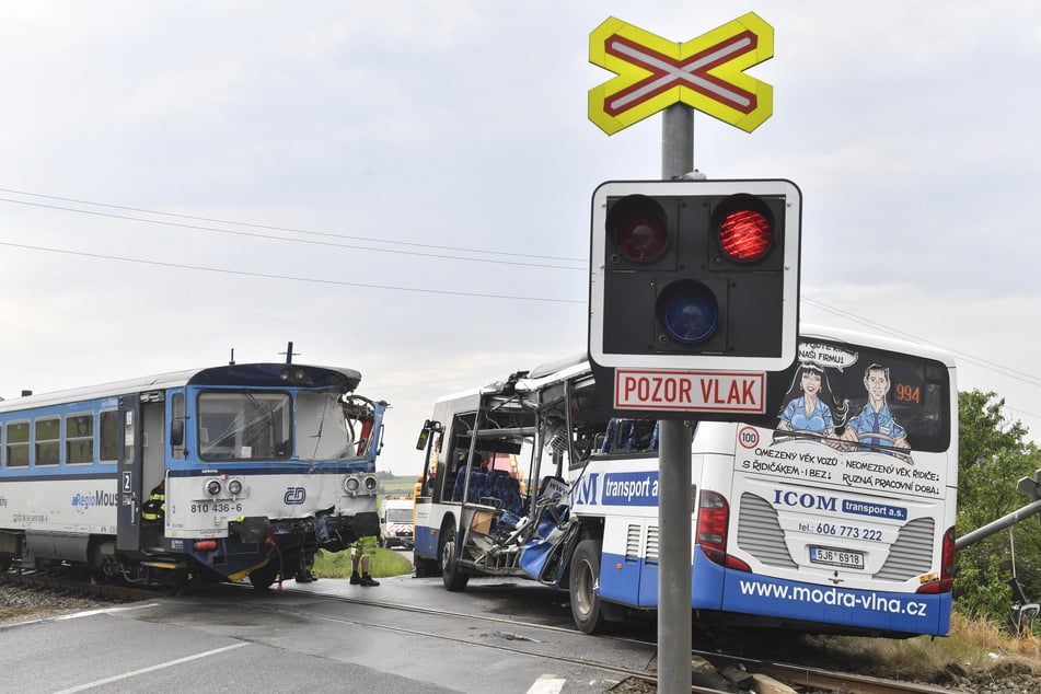 Ein beschädigter Bus und ein beschädigter Zug stehen an einem Bahnübergang. Beim Zusammenstoß eines Eisenbahntriebwagens mit einem Autobus sind in Tschechien zehn Menschen verletzt worden.