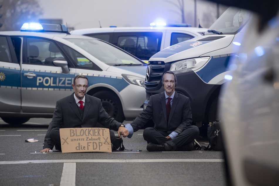 Demonstranten der Gruppe "Letzte Generation" blockieren immer wieder Straßen. Das Foto zeigt Aktivisten mit Masken des FDP-Chefs auf einer Kreuzung an der Landsberger Allee in Berlin.
